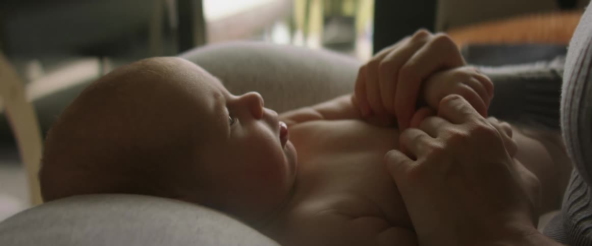 A baby is laying across their mothers lap facing up and and smiling and trying to bite their hand and the mother removes the hand from the babies mouth and holds their hands bouncing them up and down while the baby starts to close their eyes