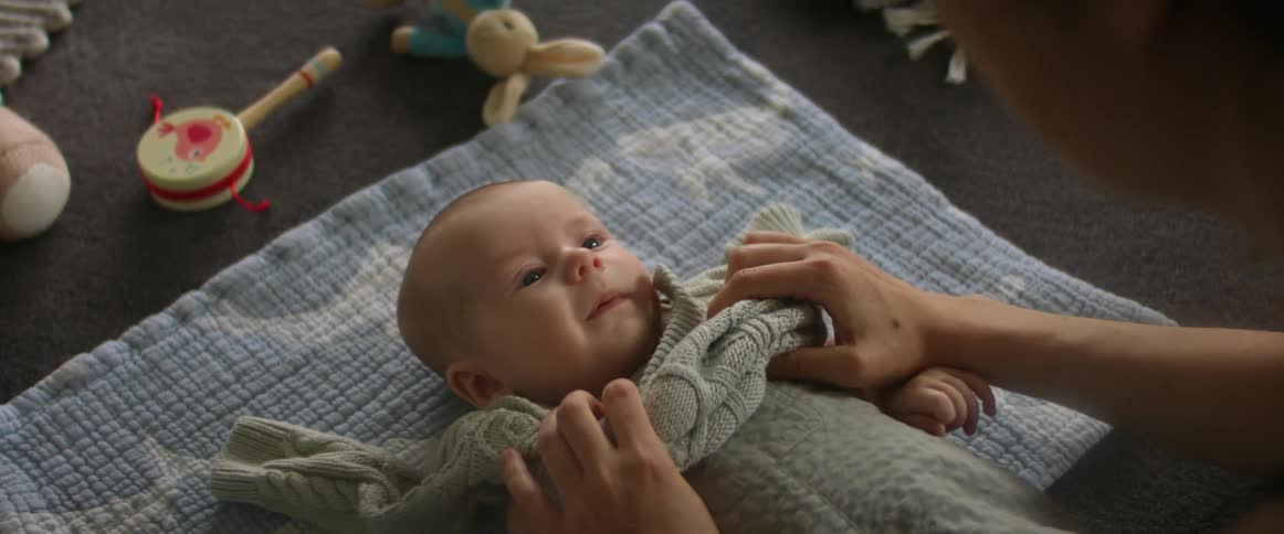 A baby lies on a blanket at home as his mother puts a sweater on him