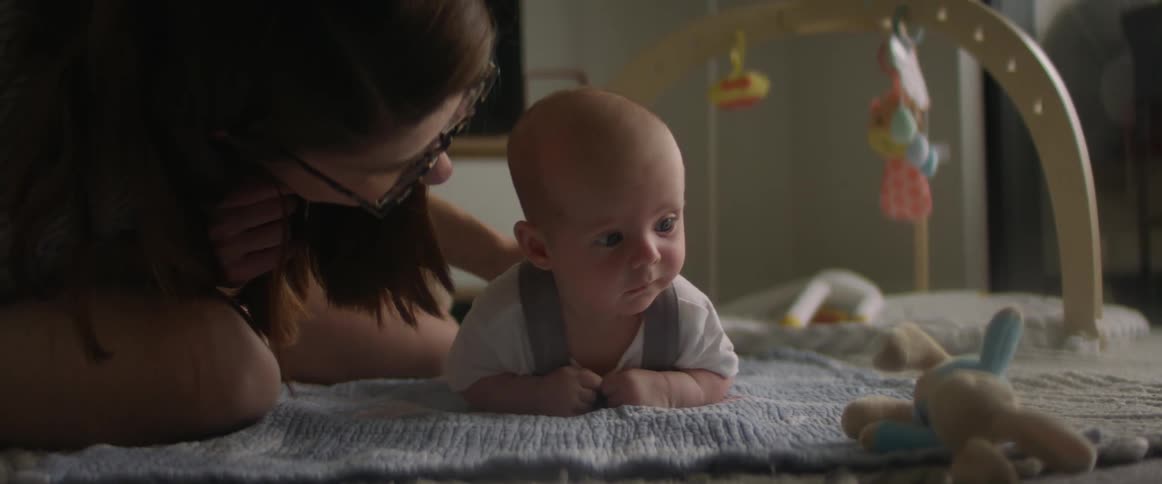 A baby is laying on his tummy on a blanket while his mother rubs his back and gives him a kiss