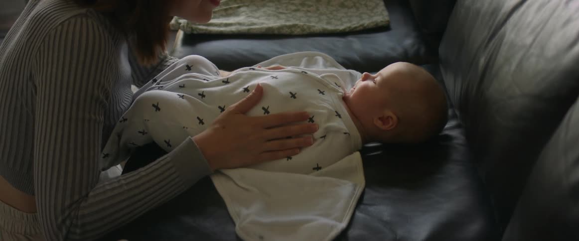 A baby lays on a couch being swaddled by his mother