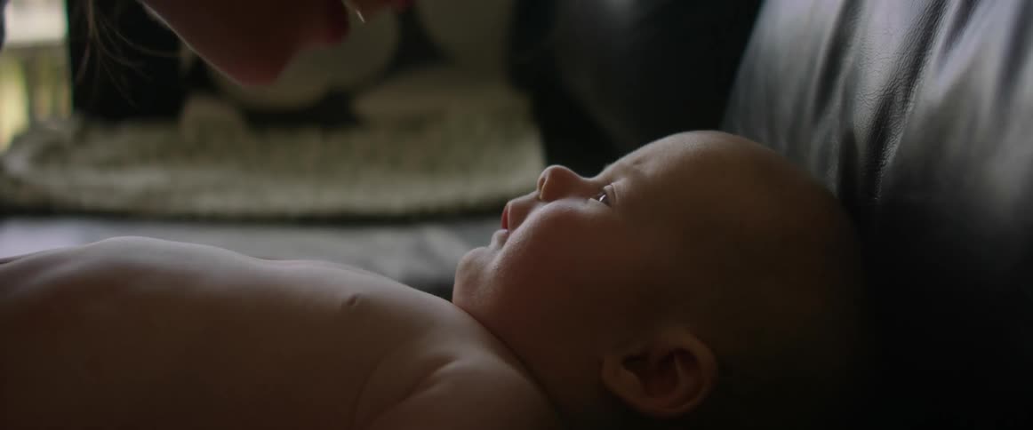 A baby lies on a couch at home and his mother bends down to kiss his nose and tummy