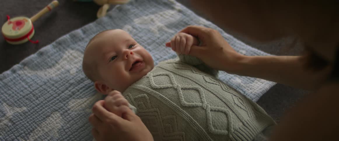A baby boy is laying on a blanket looking up and smiling at his mother who has her hands on his and is moving them up and gently waving his arms back and forth