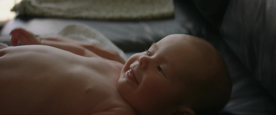A baby without clothes on laying on a couch as an adult runs their fingers over the babies belly