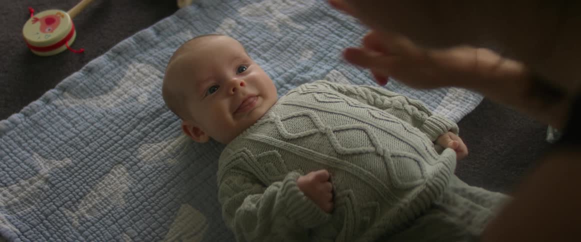 A mother is sitting leaned over her baby boy who is laying on a blue blanket and smiling up at her pulling down the sweater and touching his nose with her finger