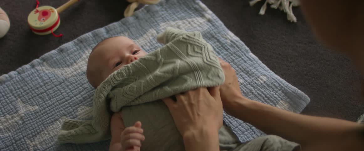 A woman is putting a sweater on her baby as they lay on a blanket