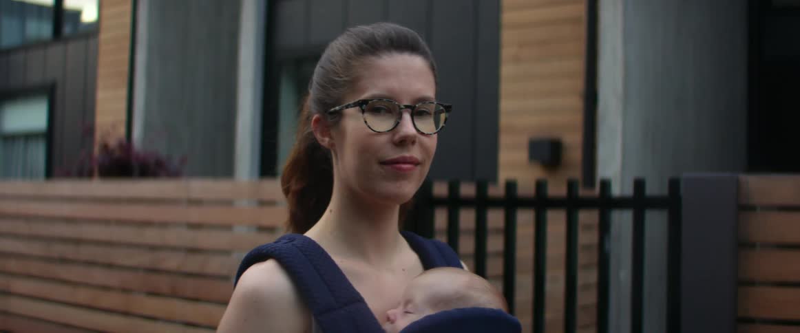 A mother stands outside a building with her infant son in a baby carrier on her frontside