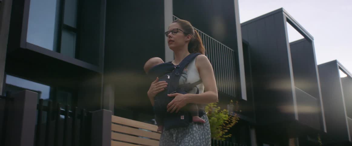A mother has her baby in a baby sling on her chest as she walks by some apartment buildings outside