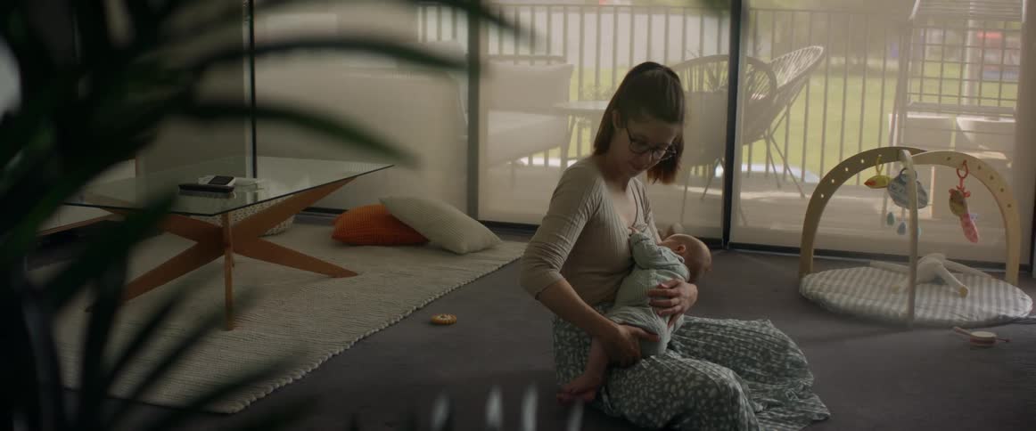 Mother with ponytail glasses brown sweater and skirt breastfeeds baby in the living room at home