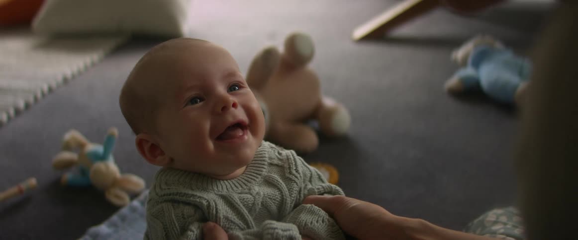 An adult holding a baby up right on the floor as the baby smiles