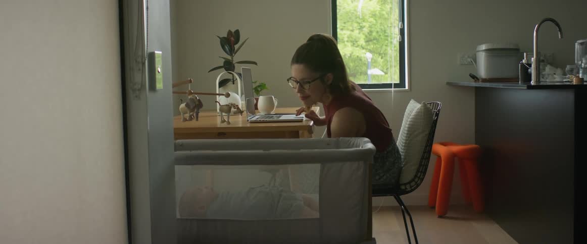 A woman is sitting at a table typing on a laptop computer as she reaches down to comfort her baby sleeping in a playpen next to the table