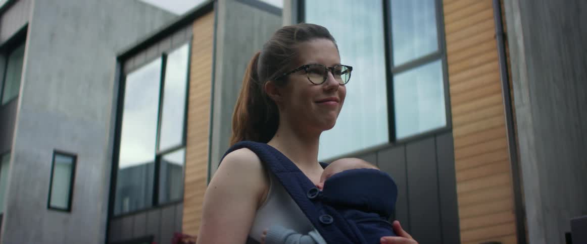 A mother is standing outside an apartment building while smiling and rocking her baby