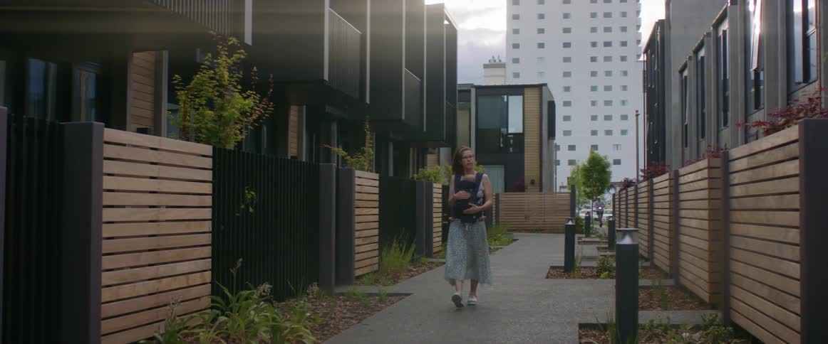 A woman carries a baby while walking along a walkway between two buildings in a city during the day