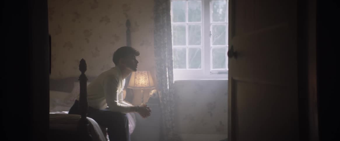Man leans forward while sitting on bed in dimly lit bedroom