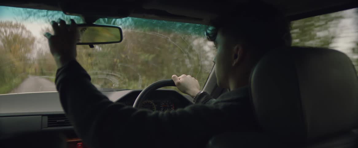 A teenage boy is driving an automobile on a country road lined with trees as he adjusts his rear view mirror