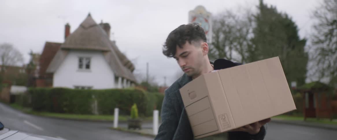 A man is carrying a box to a car on a neighborhood street and he puts it in the trunk