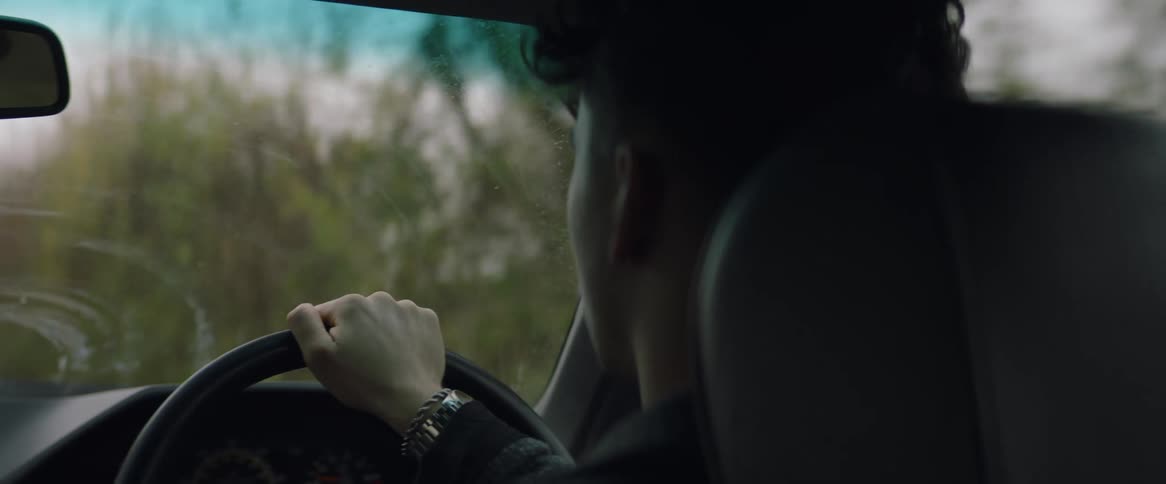 A man is driving an automobile and holding the steering wheel with one hand as he travels on a country road lined with trees on a fall day