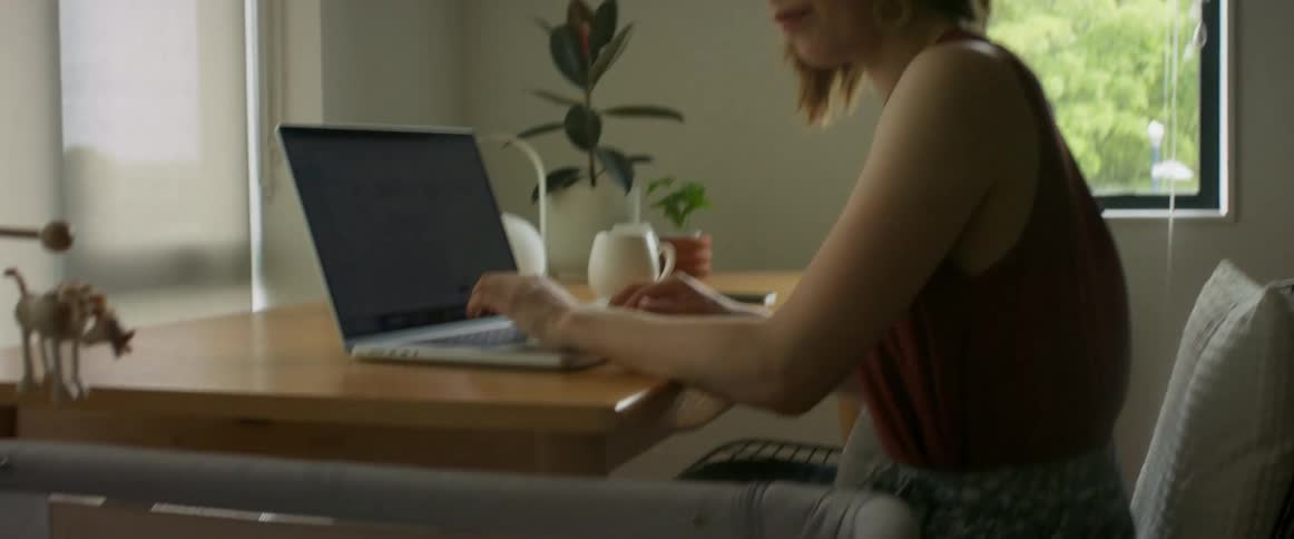 A woman is sitting at a table typing on a laptop computer as she looks at her baby sleeping in a playpen next to the table
