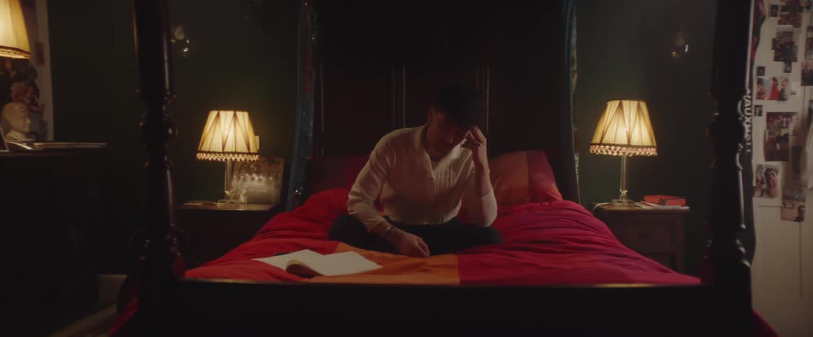A man is sitting on his bed with an open book and his head resting on his hand
