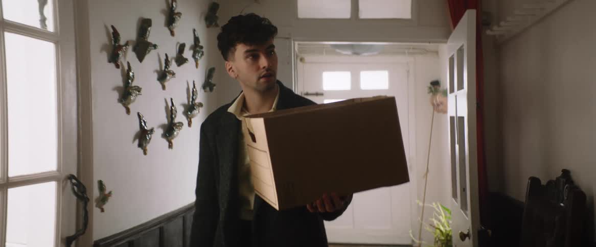 A man carrying a box pushes a door open and walks into the room looking around