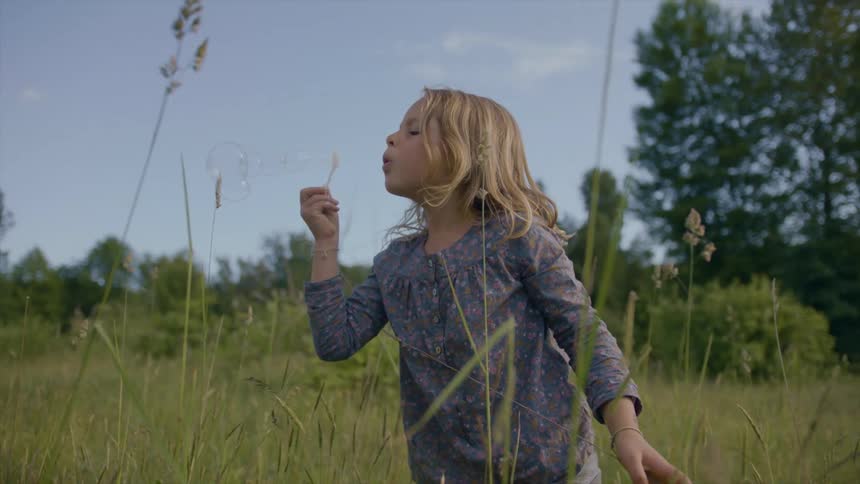 A little girl with long blond hair is standing outside in the weeds blowing soap bubbles
