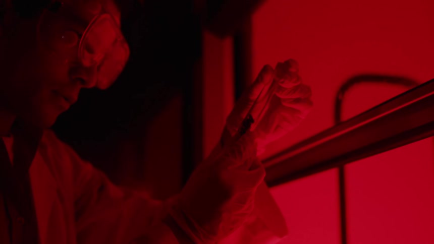 A man wearing goggles is standing in room illuminated with red light looking at a device in his hands