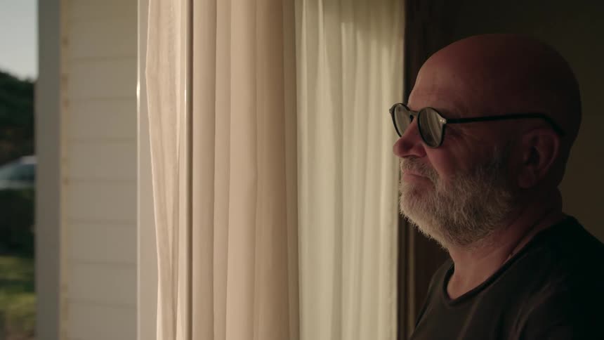 A man is standing inside his home as he opens the curtain and looks outside