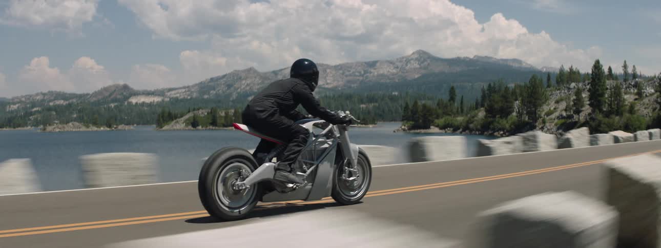 A motorcycle travels across a bridge toward a forested mountain as the rider is wearing a black jacket and helmet