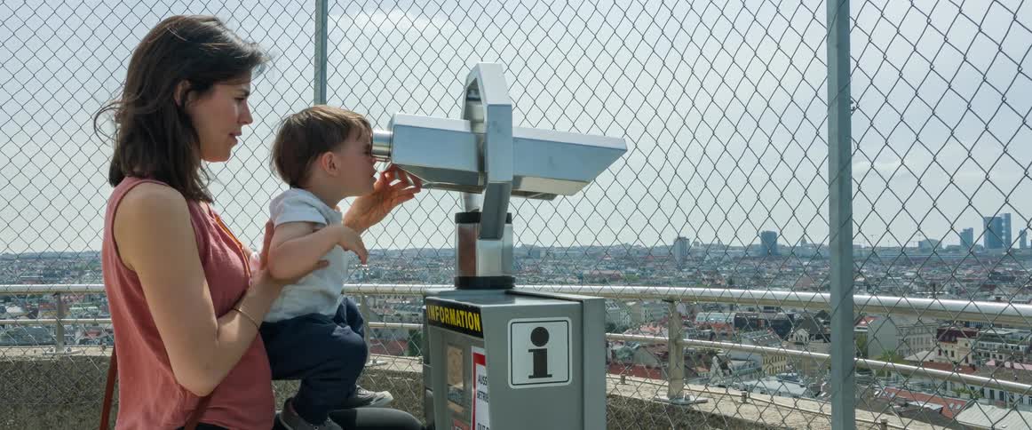 A woman is standing on a rooftop with her young son as she holds him while he looks through a telescope at the city buildings
