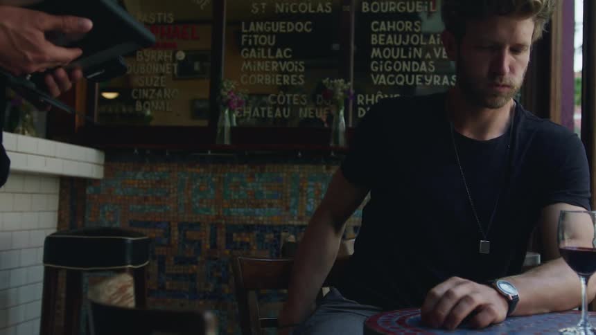A man sitting at a cafe table with wine in front of him paying the waiter and getting out his wallet