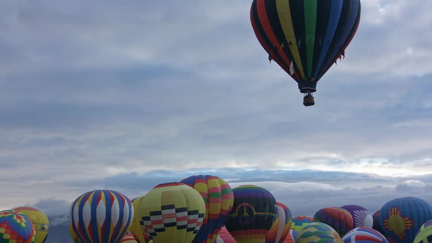 Hot air balloons rise into air above other hot air balloons clustered together on ground