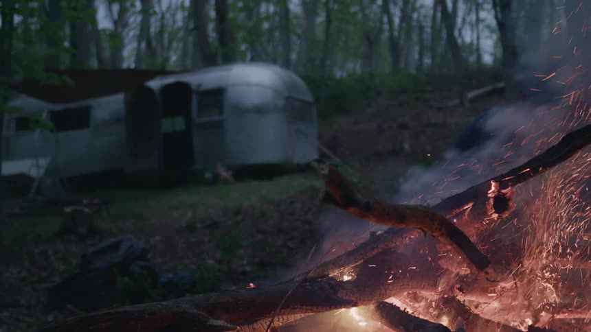A bonfire up close and a man throwing debris onto it with a camper in the background