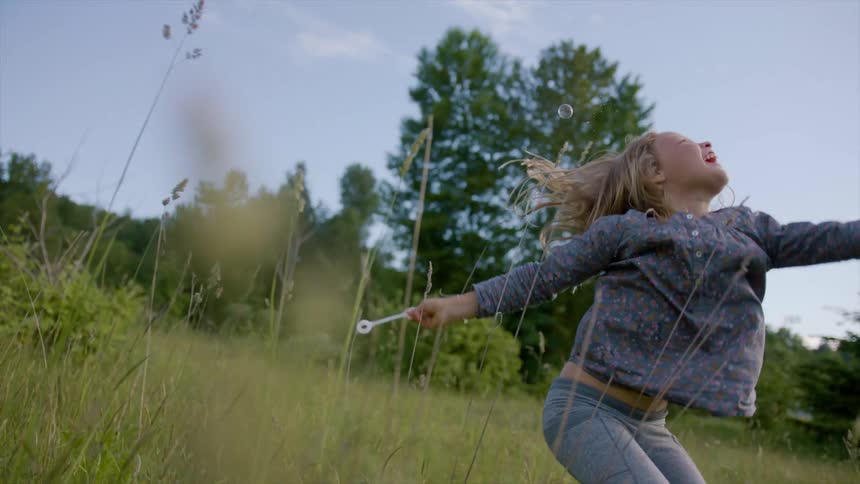 A young girl in a field playfully jumps up to try to pop a bubble that she blew