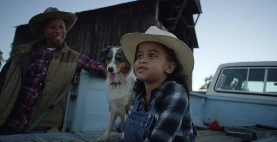 A little girl wearing a cowgirl hat sits on the back of a truck next to a dog as a man stands next to them smiling and looking at the girl