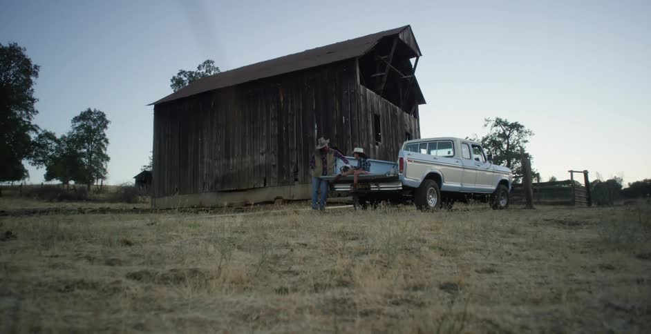 Adult man leans against truck as girl sits in truck bed at farm