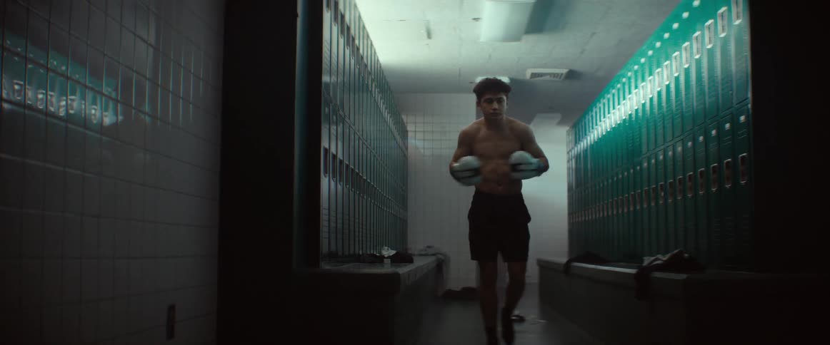 A teenage boy is standing in a locker room wearing boxing gloves and doing arm exercises and movements as he bounces up and down