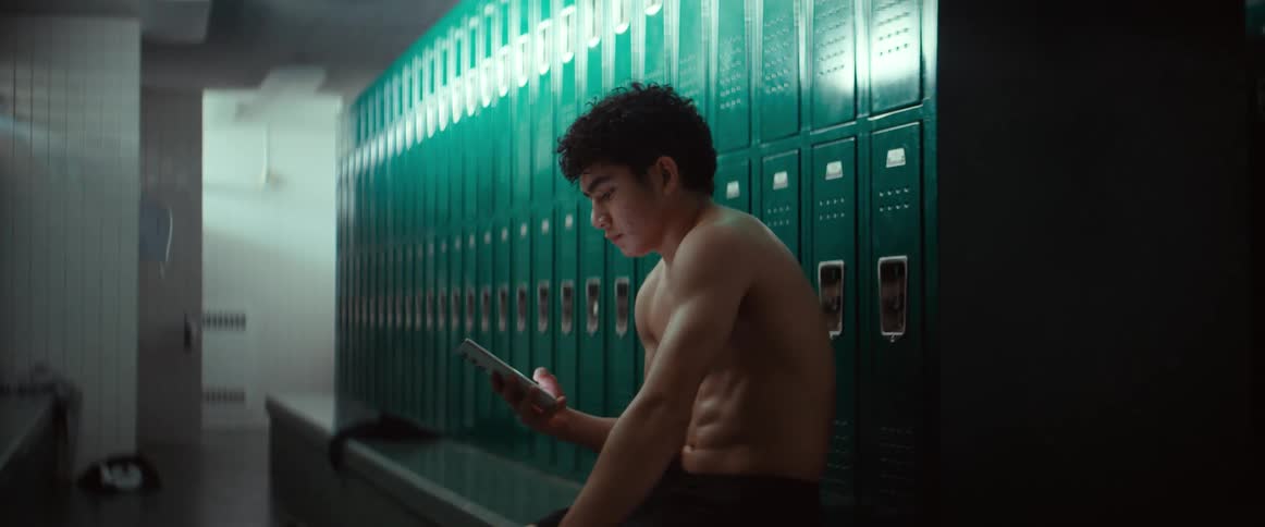 A muscular young male sits alone with his phone while in a locker room