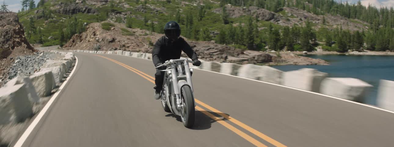 Motorcyclist rides along highway crossing water with mountains in the background