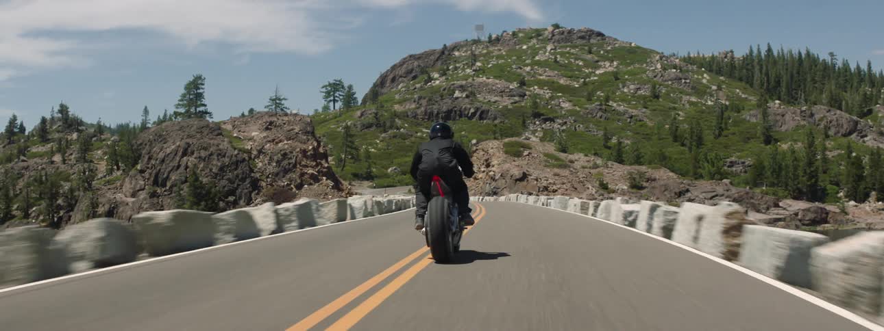 A motorcycle travels across a bridge toward a forested mountain as the rider is wearing a black jacket and helmet