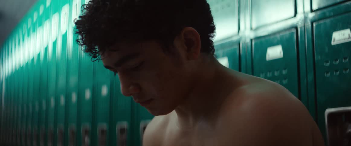 A bare chested man is sitting in a locker room holding his phone with both hands as he texts with his thumbs