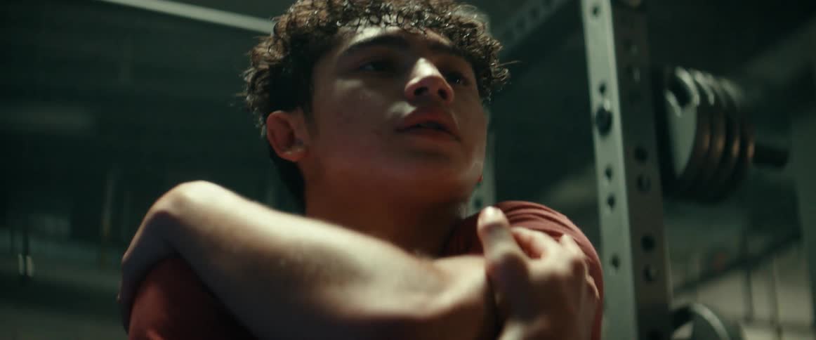 A young man is stretching his arms in a gym while surrounded by weights