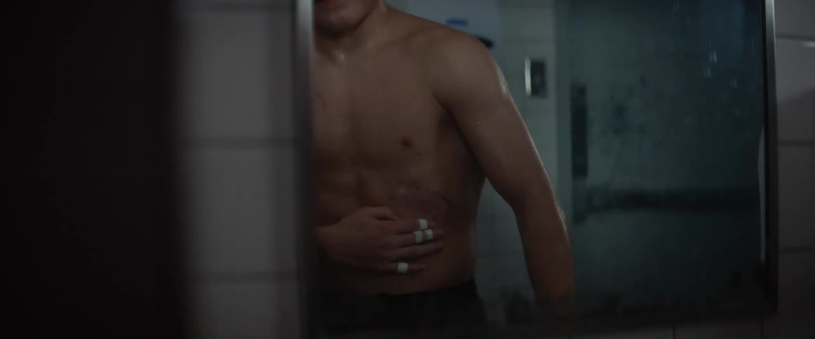 A man shirtless stands in front of a mirror looking at his body and holding his stomach in pain