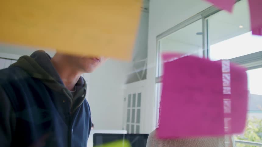 A teenage boy and girl are standing together as they put colored note on glass in a room