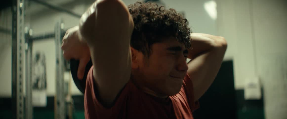 A teenage boy is working out in the gym lifting a weight over his head
