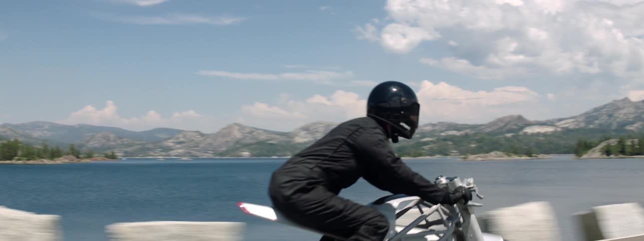 Motorcyclist rides along highway across lake from mountains and forests ona cloudy day