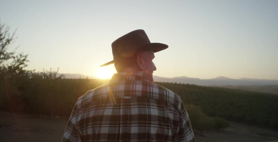 A man wearing a cowboy hat and a plaid flannel shirt is standing outside as the sun shines brightly over the mountains in the distance