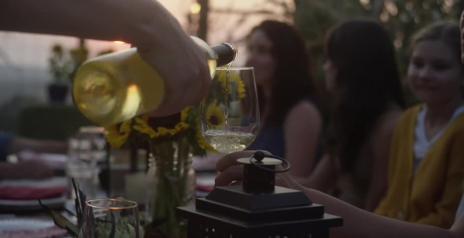 Friends sitting at outdoor table watch as person fills wine under a red sky