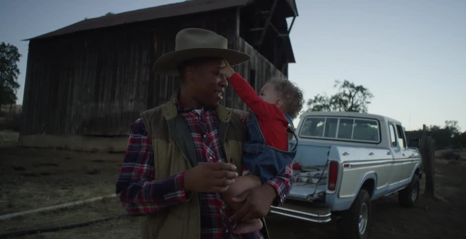 A man holds his son while standing by their pickup truck next to a barn
