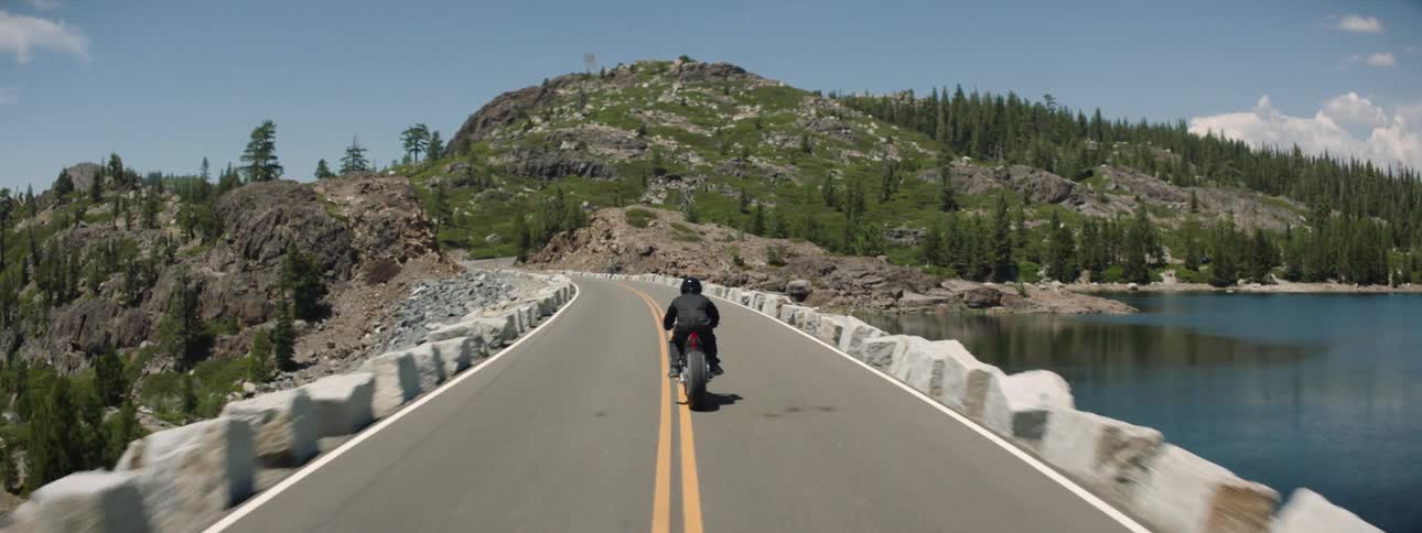 A person rides a motorcycle across a bridge heading toward a mountain range