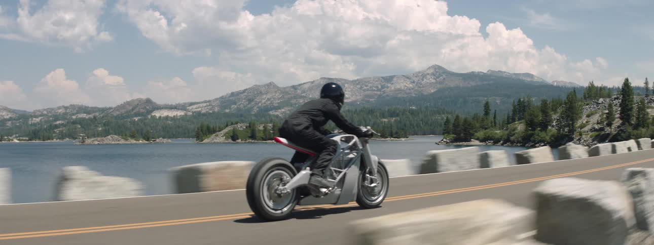 A person driving a motorcycle down a road with a lake and mountains in the background