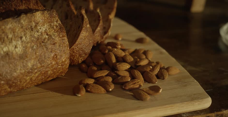 Sliced hearty bread and almonds sit on large wooden chopping board on wooden dining table